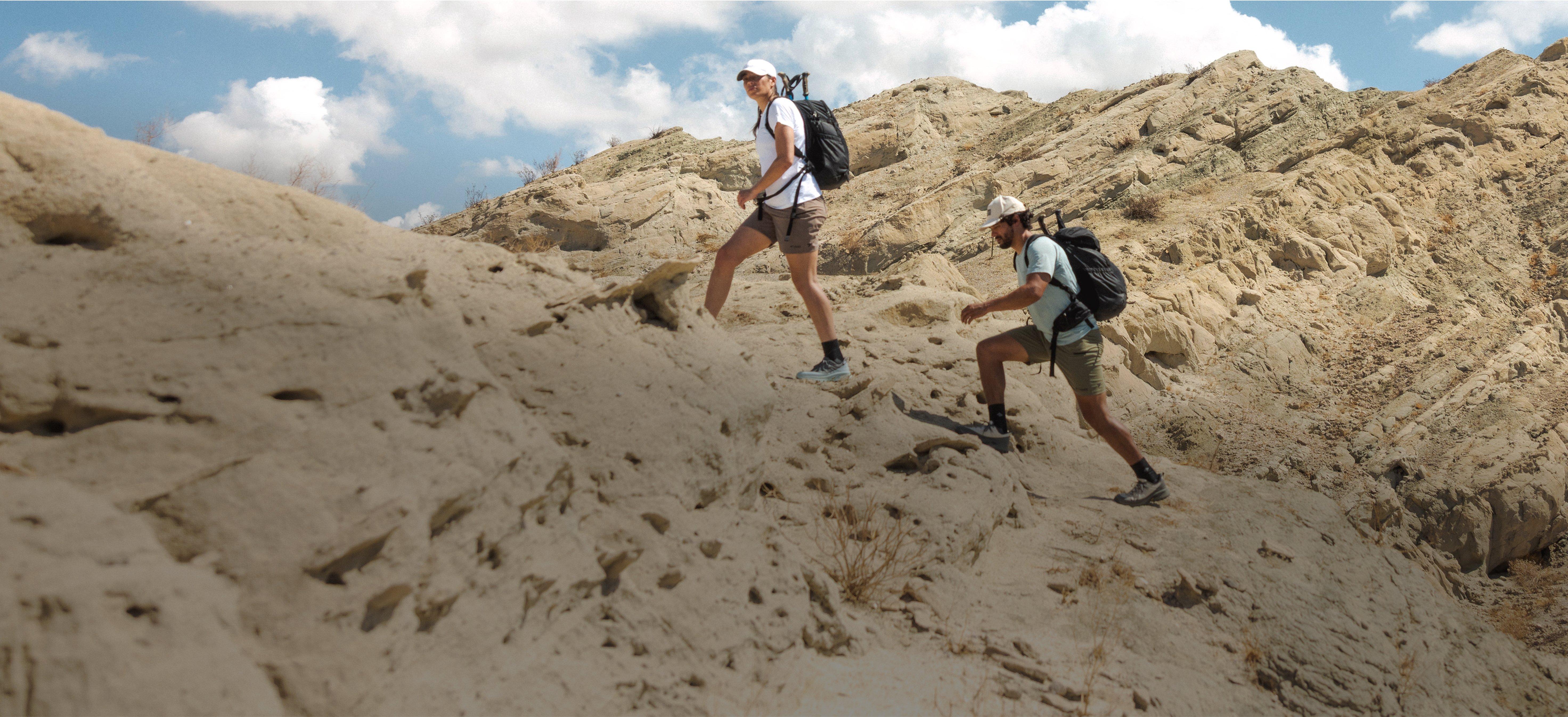 Two hikers with backpacks trekking up a rocky desert trail under a blue sky with clouds.