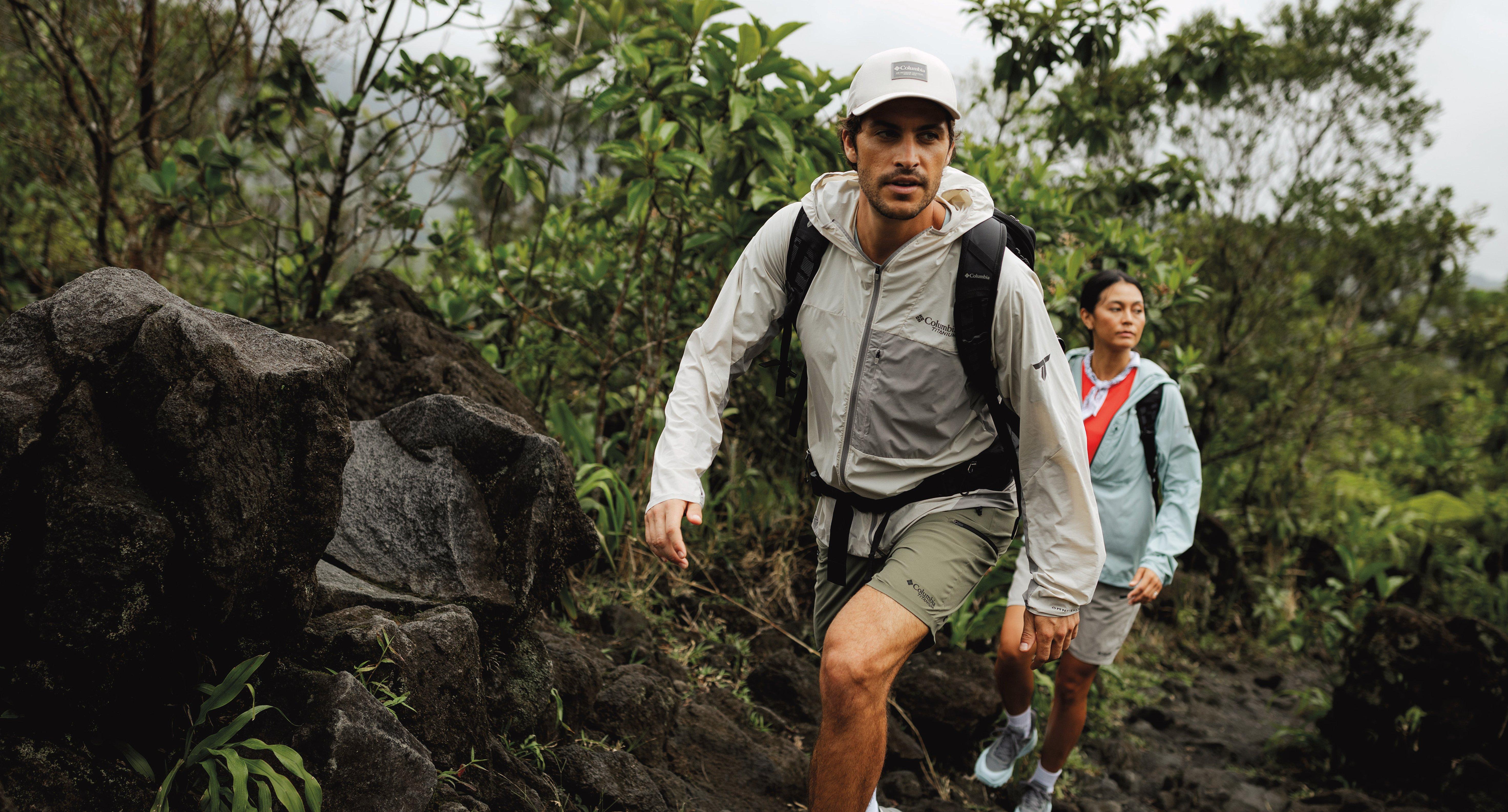 Two hikers wearing jackets and backpacks trek through rocky terrain surrounded by lush green vegetation.