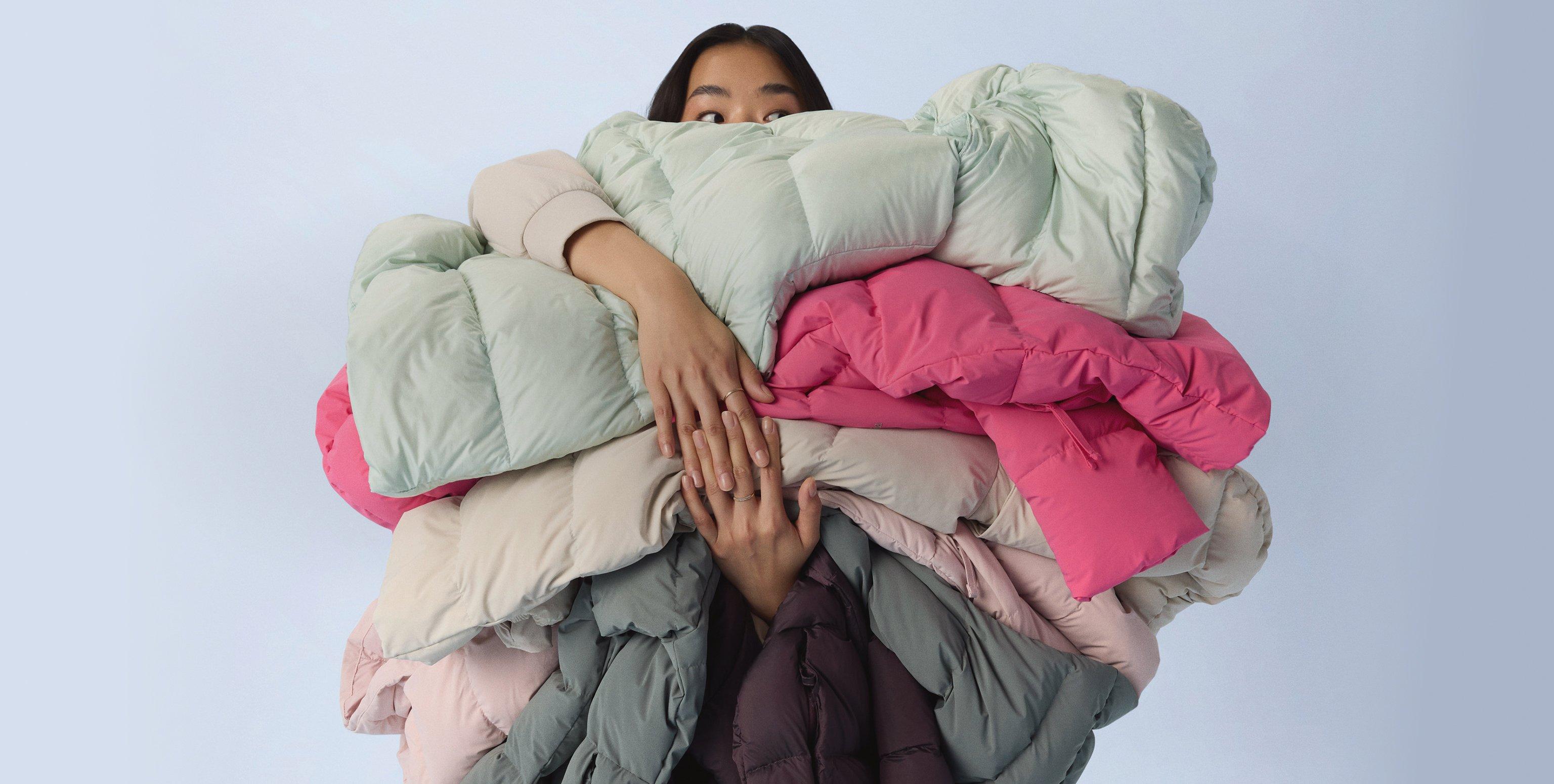 Person holding a large armful of colorful puffer jackets in mint, pink, beige, and gray tones.