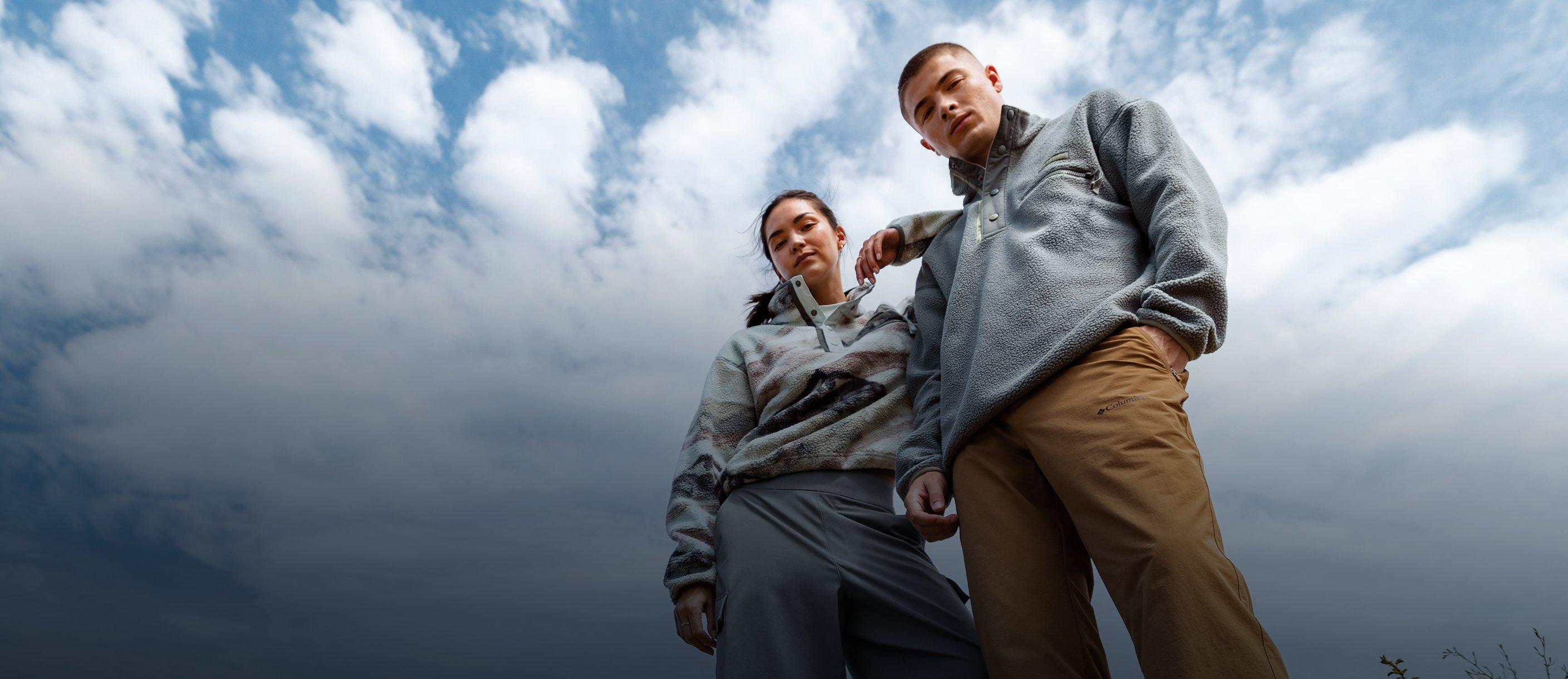 Two people standing outdoors wearing fleece jackets and casual pants against a cloudy sky.