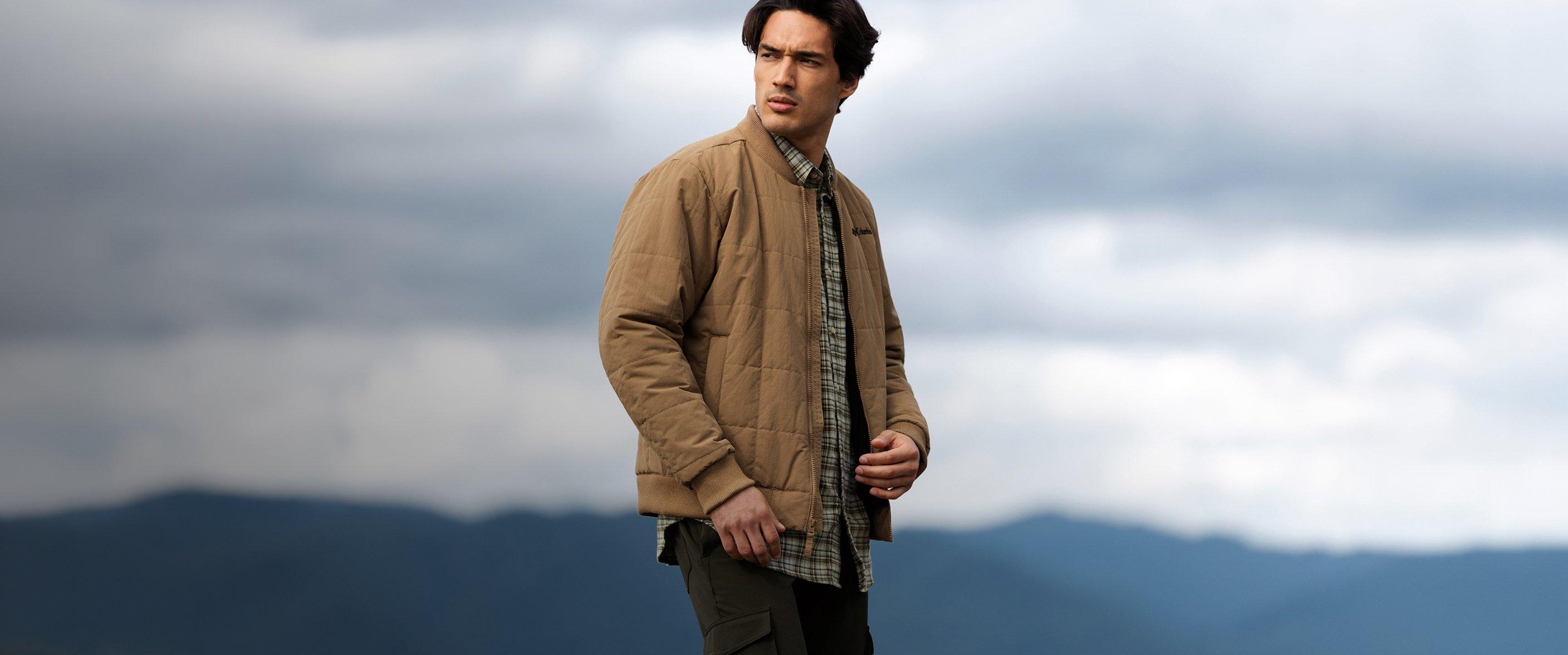 Man wearing tan quilted jacket over plaid shirt and olive cargo pants, standing outdoors.