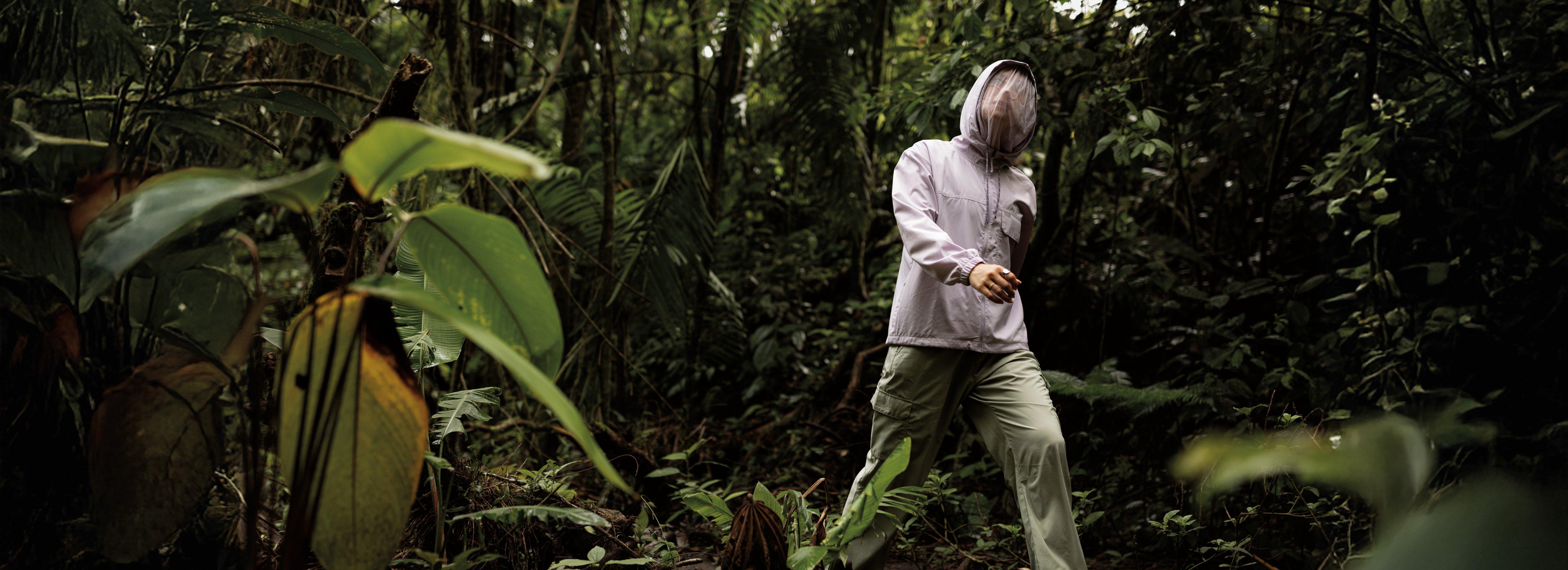 A man standing in a jungle with a white jacket on with the hood up, showcasing the netting around his face for insect protection.