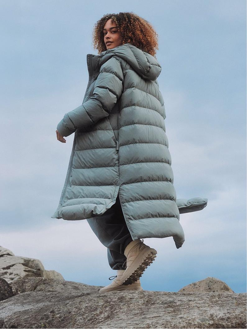 Woman walking on rocks wearing a sage green quilted puffer coat with hood, black pants, and tan boots.