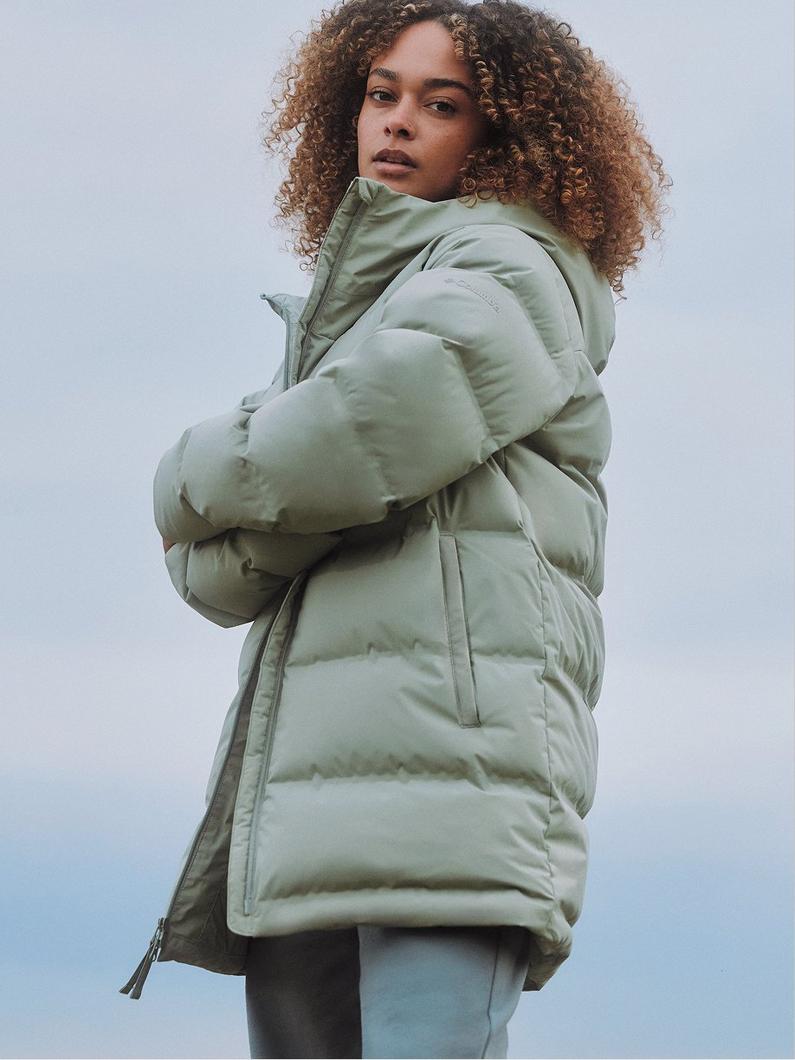 Model wearing a sage green quilted puffer jacket with high collar and side pockets, arms crossed.