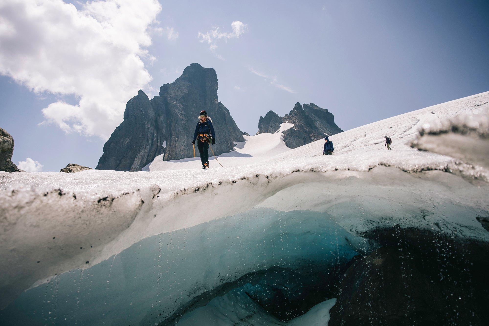 Three mountaineers traverse a snow-covered glacier beneath a rugged mountain peak under a partly cloudy sky. The image captures a dramatic perspective, showing a translucent blue crevasse beneath the icy surface. The climbers are roped together and equipped with helmets, harnesses, and crampons, highlighting the technical nature of their alpine journey.