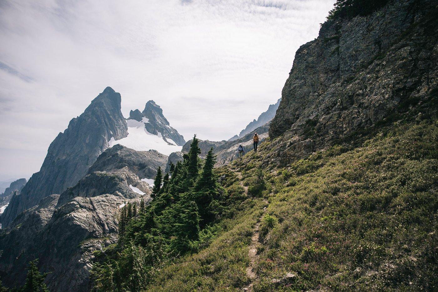 Two hikers navigate a narrow alpine trail along a steep, rocky slope surrounded by rugged peaks and scattered snowfields. In the distance, jagged mountains tower above a dramatic landscape of cliffs and evergreen trees. The terrain ahead appears challenging and exposed.