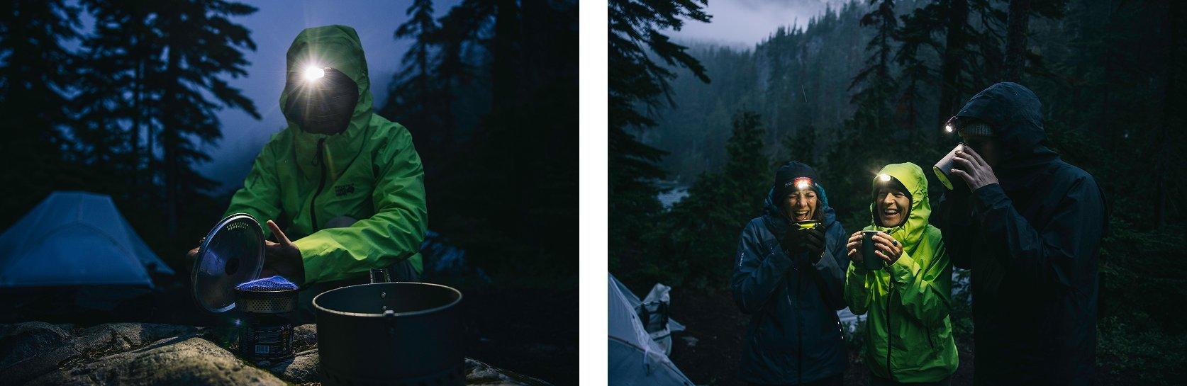 In a misty forest campsite at dawn or dusk, a person in a bright green rain jacket prepares a meal on a camping stove, illuminated by a headlamp. In the adjacent image, three campers wearing headlamps and rain jackets laugh and sip hot drinks together, enjoying a cozy moment in the cold, damp wilderness. Tents and tall evergreen trees surround the group, with a faint view of a lake and mountains in the background.