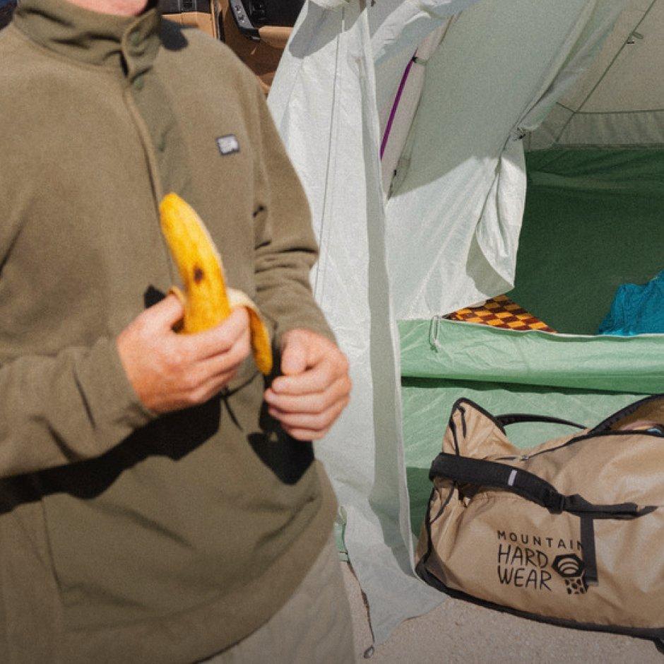 An unzipped Camp 4™ Duffel 65 sits in the dirt outside an open tent. In the foreground a man eats a banana.