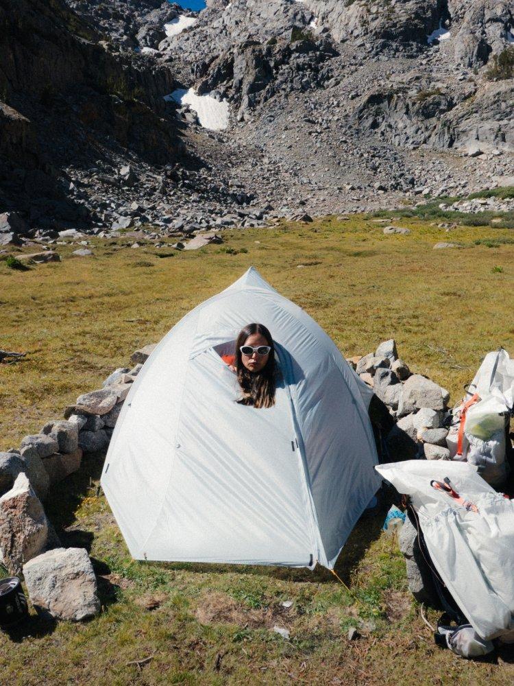 A woman sassily pops her head out of a mostly zipped-shut Nimbus™ UL 2 Person Tent with loaded packs outside.