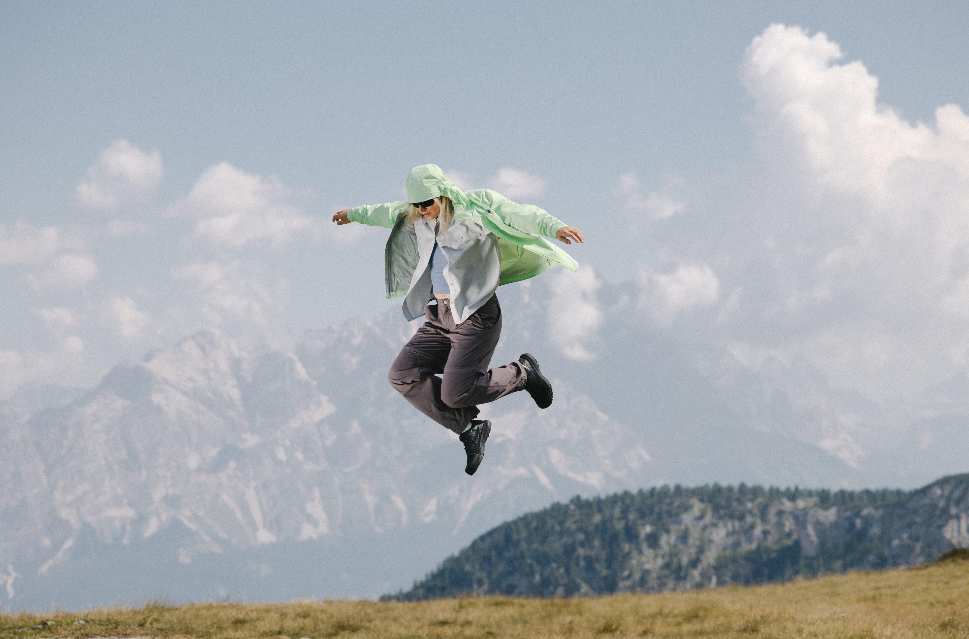 Floating mid-leap, a woman’s unzipped Cloud Cipher™ Hooded Jacket floats like a cape as she extends her arms like wings.
