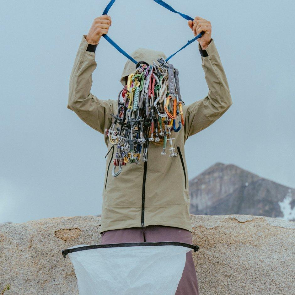 A person wearing a Chockstone™ Alpine Light Hooded Jacket hoists a sling full of cams and carabiners with a snowy peak in the background.


