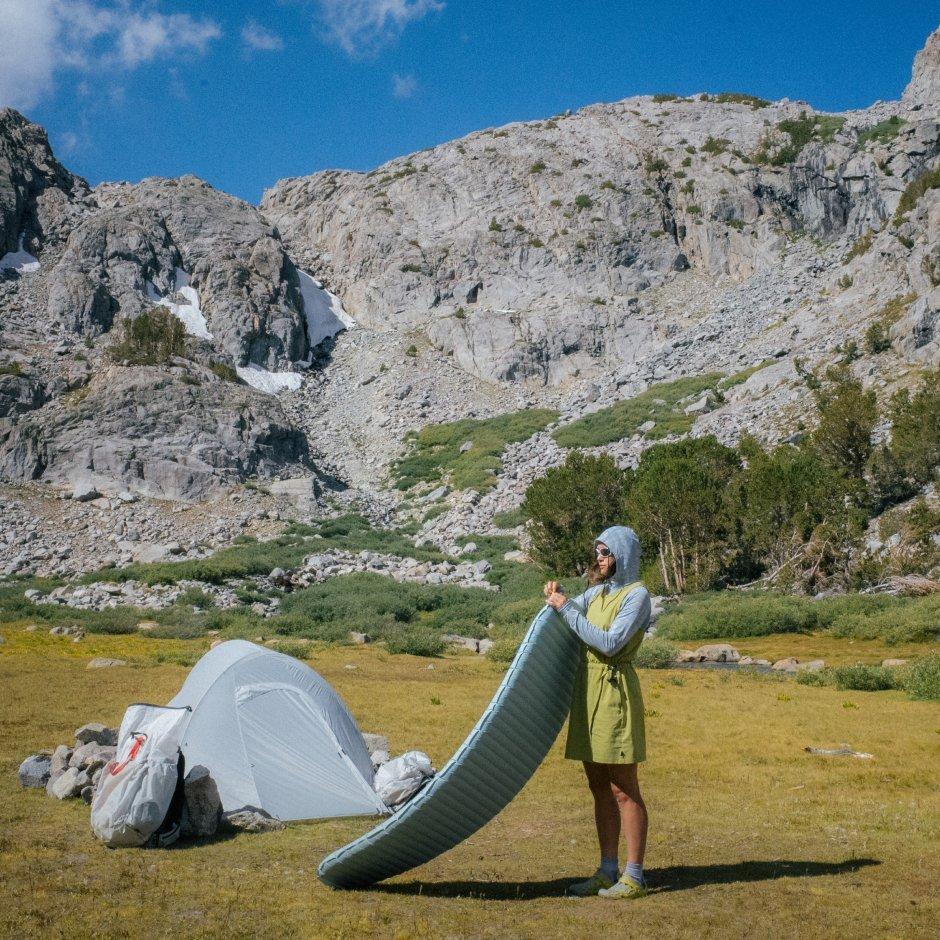 Woman in a Crater Lake™ Long Sleeve and Dynama™ Dress sets up a tent and mat in a clearing at the foot of the mountain.