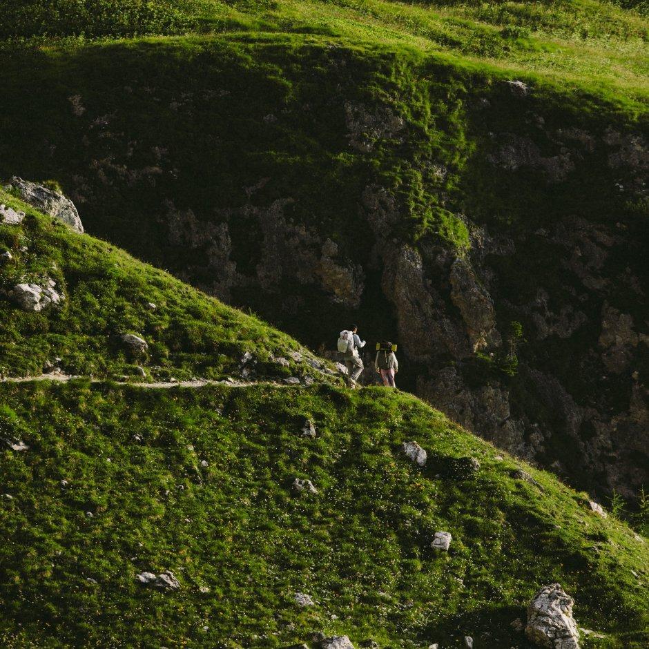 In the distance, amidst greenery, two people with backpacking gear and hike poles begin to descend out of view. 