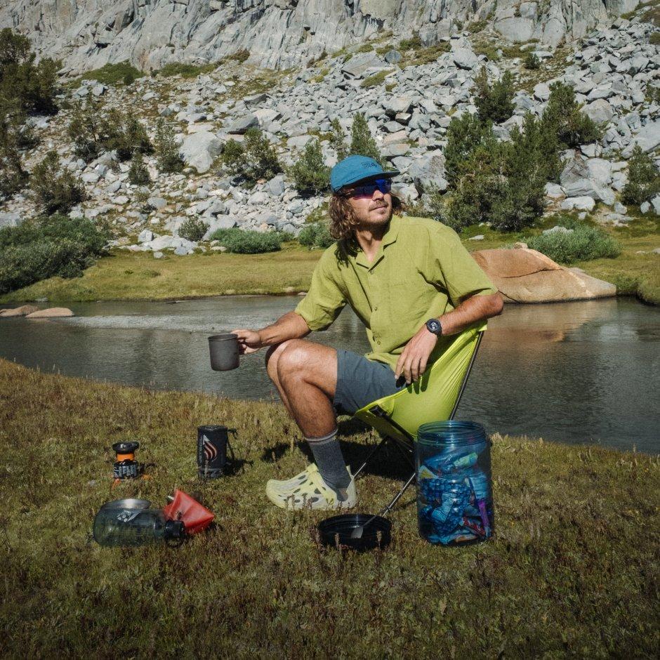 A man in a Sun Drift™ Cooling Short Sleeve Shirt and Rockrydge™ Shorts sits by a bear canister, lakeside.