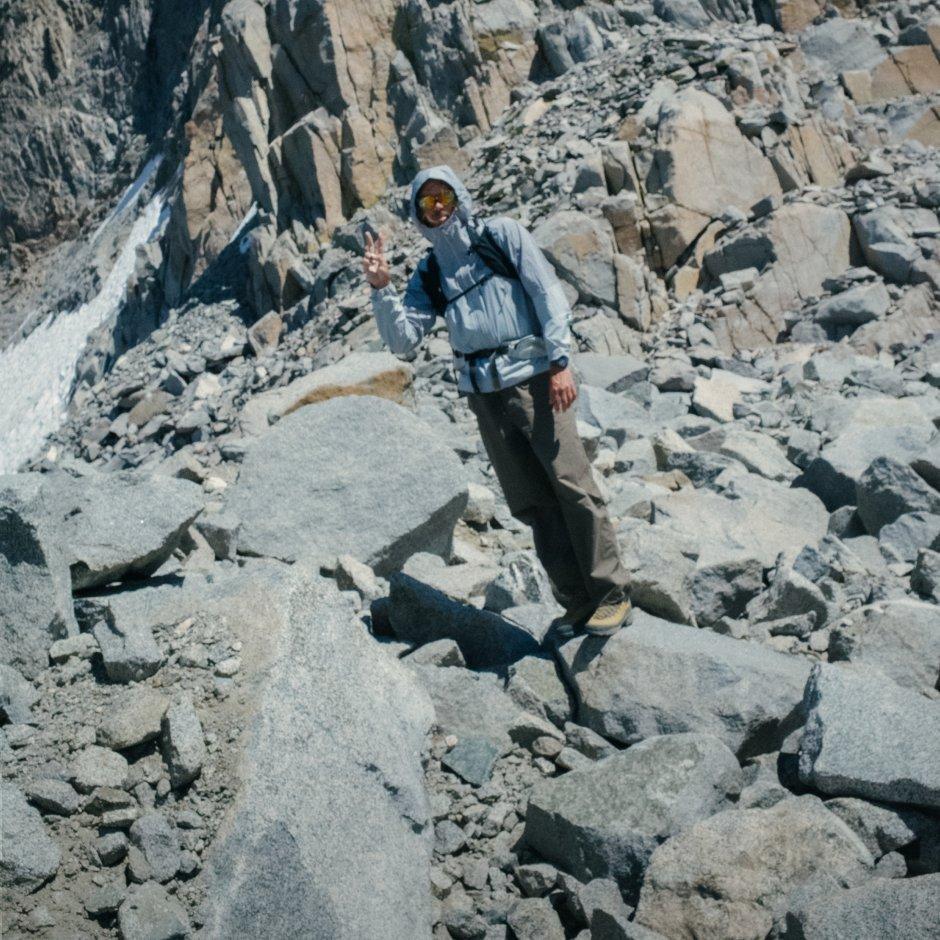 Wearing a Stryder™ Jacket with the hood up, a backpacking man holds up a peace sign poses on rocky terrain.