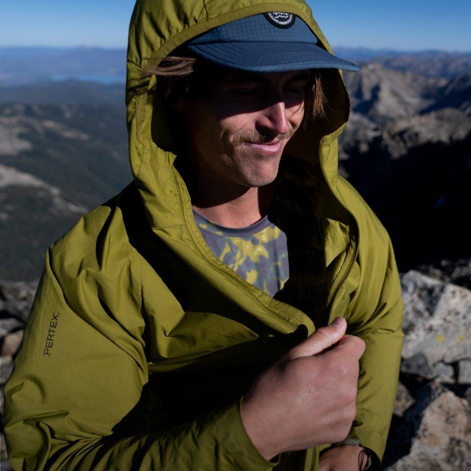 Atop the windy summit, a mustached man zips up his Kor AirShell™ Hoody over his baseball hat.


