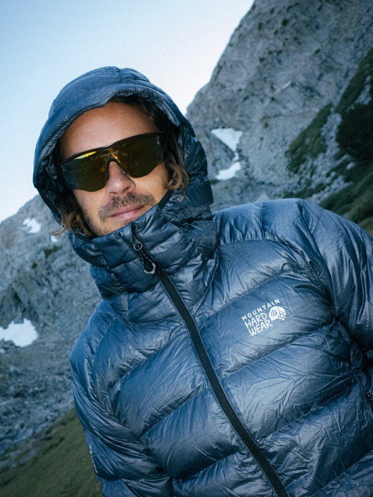 Mustached man in sunglasses and Phantom™ Alpine Down Hooded Jacket in front of a snow-dabbed range.
