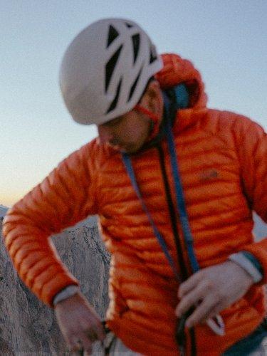 A man wearing a Ghost Whisperer™ Jacket and climbing helmet with rocky peaks in the background.