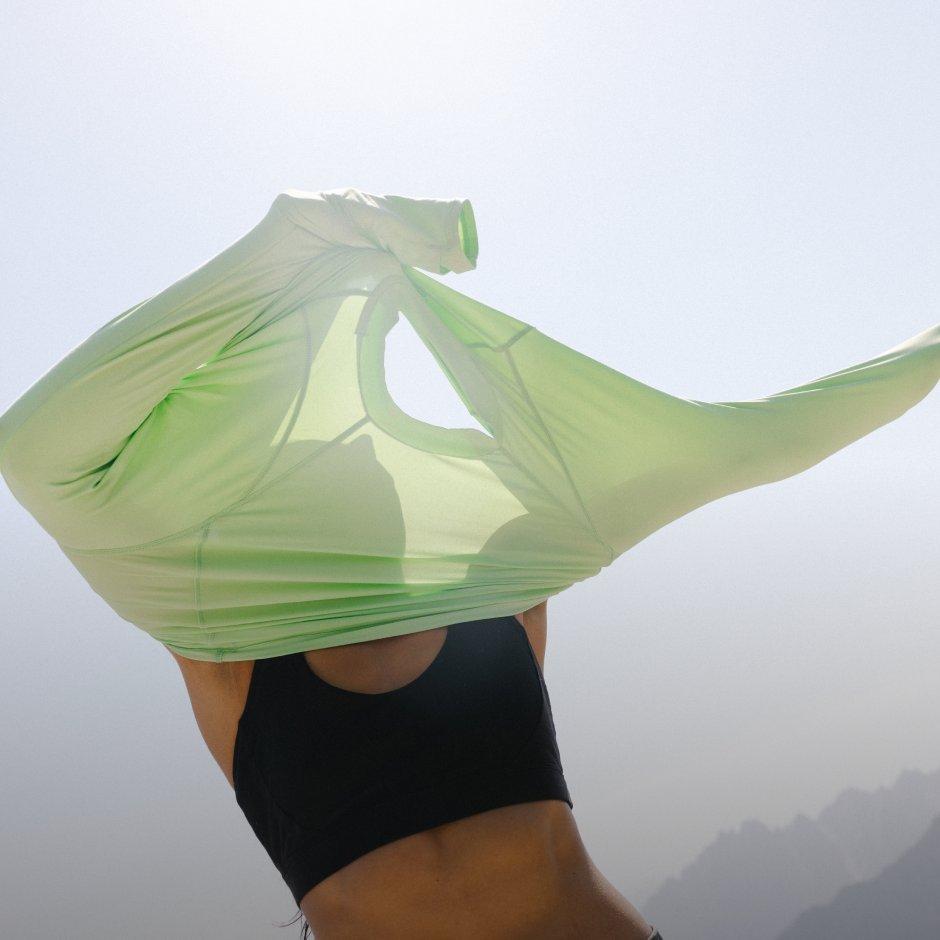 Woman in a sports bra pulling on a Crater Lake Long Sleeve Shirt. Light can be seen through the lightweight fabric.


