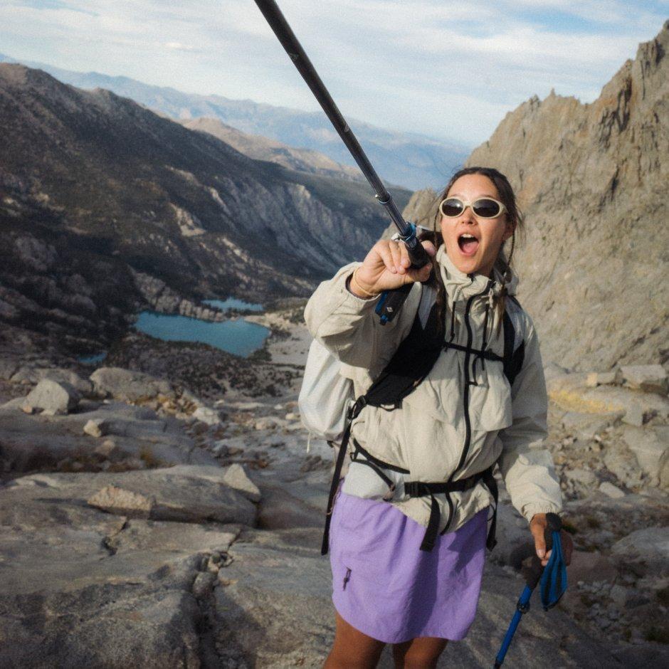 A woman with hiking pack wearing a Stryder™ Jacket and Dynama™ Skort excitedly points with her hike pole on rocky terrain.