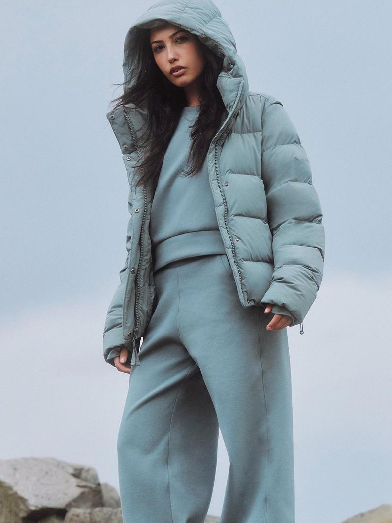 Woman wearing sage green puffer jacket with hood, matching sweatshirt and joggers in coordinated outfit.