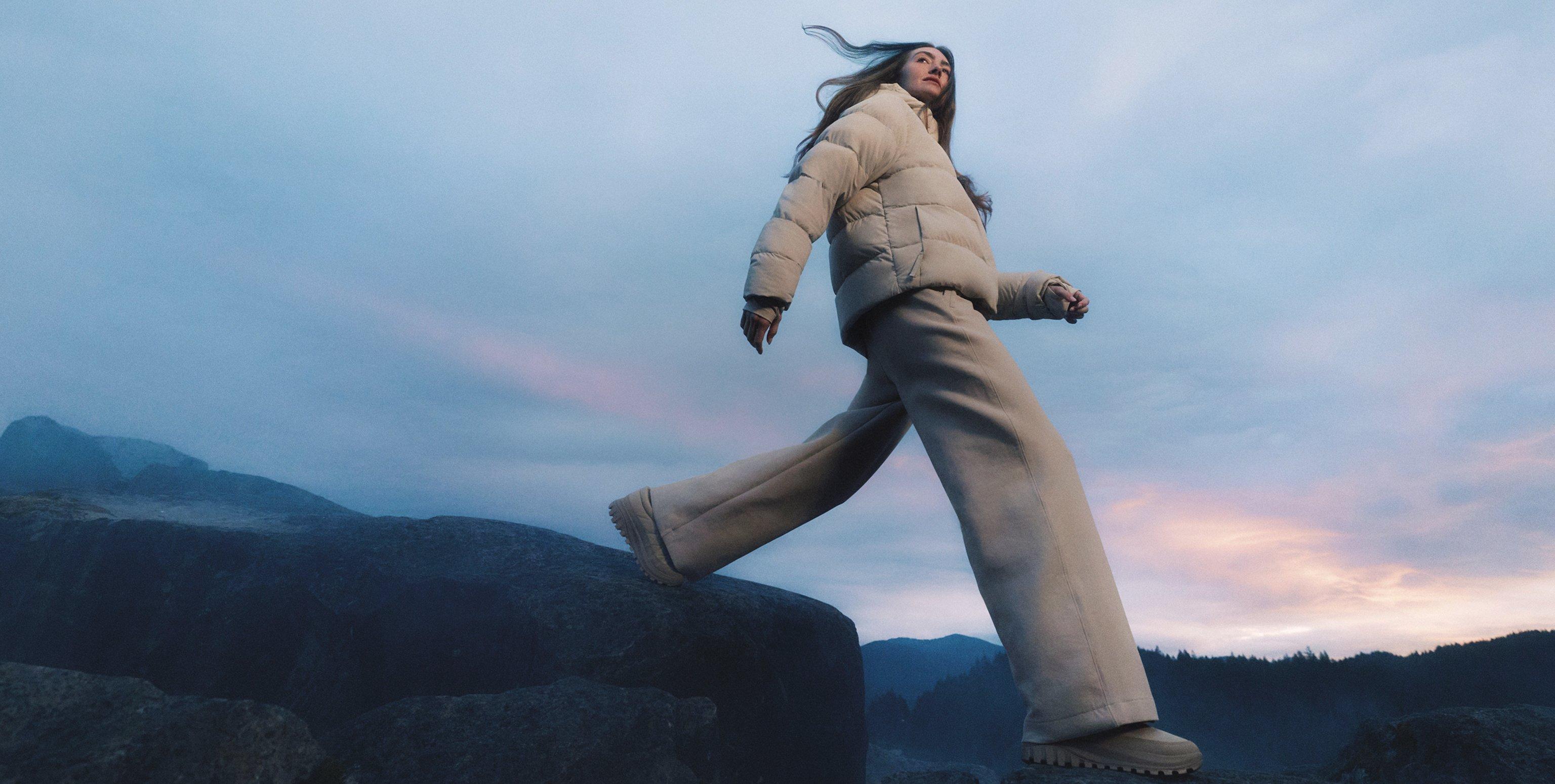 Woman jumping outdoors at dusk wearing beige puffer jacket, matching pants, and boots.