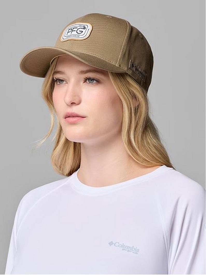 Woman wearing a tan trucker hat with circular logo patch and white long-sleeve shirt.