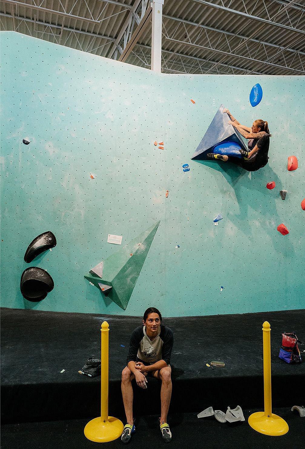 Kyra sits on the ground taking a break while Alex is near the top of a bouldering route behind her.