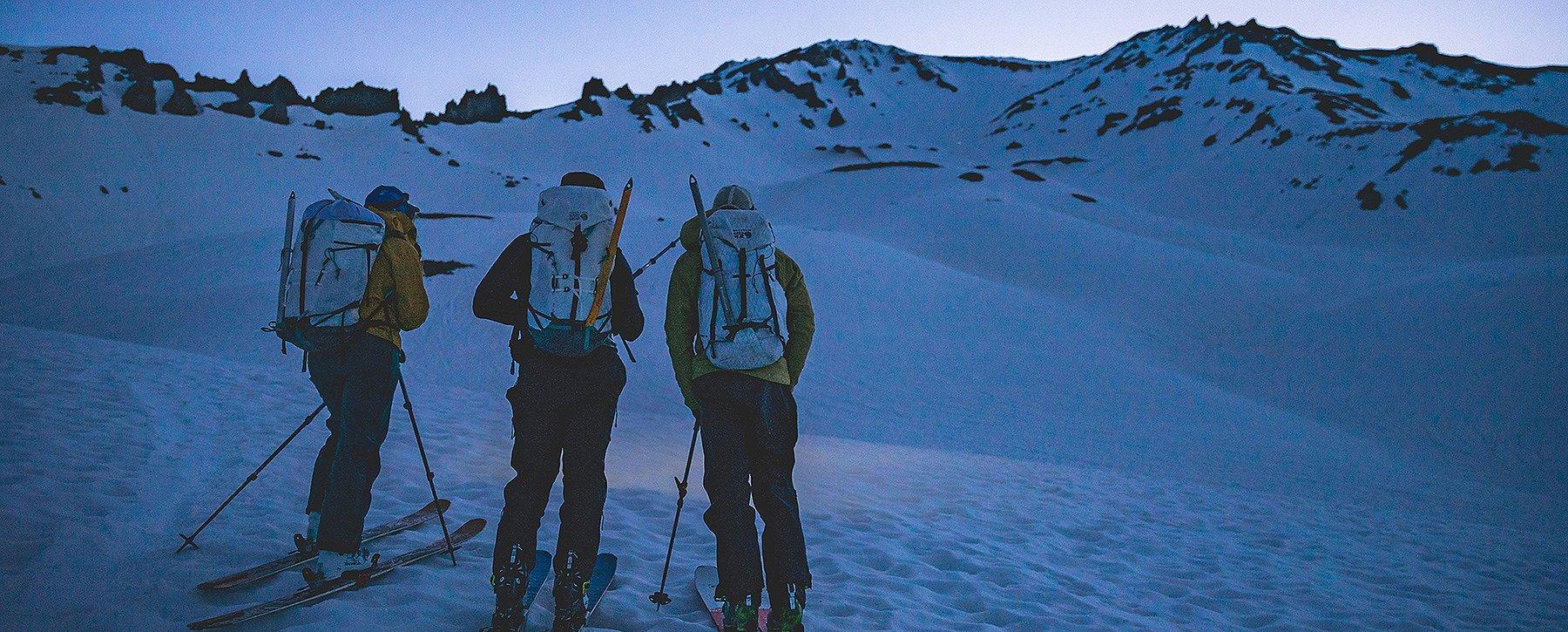 Three skiers planning their approach at dusk, looking at the mountain ahead 