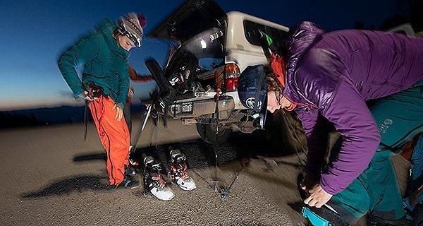 Blurry mid motion image of three skiers getting ready at the back of their truck, putting on ski boots and bibs at dusk.