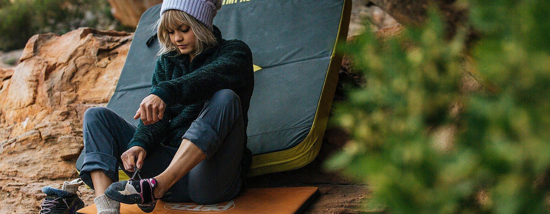 A woman putting on her climbing shoes, sitting on a crash pad, in the Women's Women's Monkey Fleece™ Pullover. 
